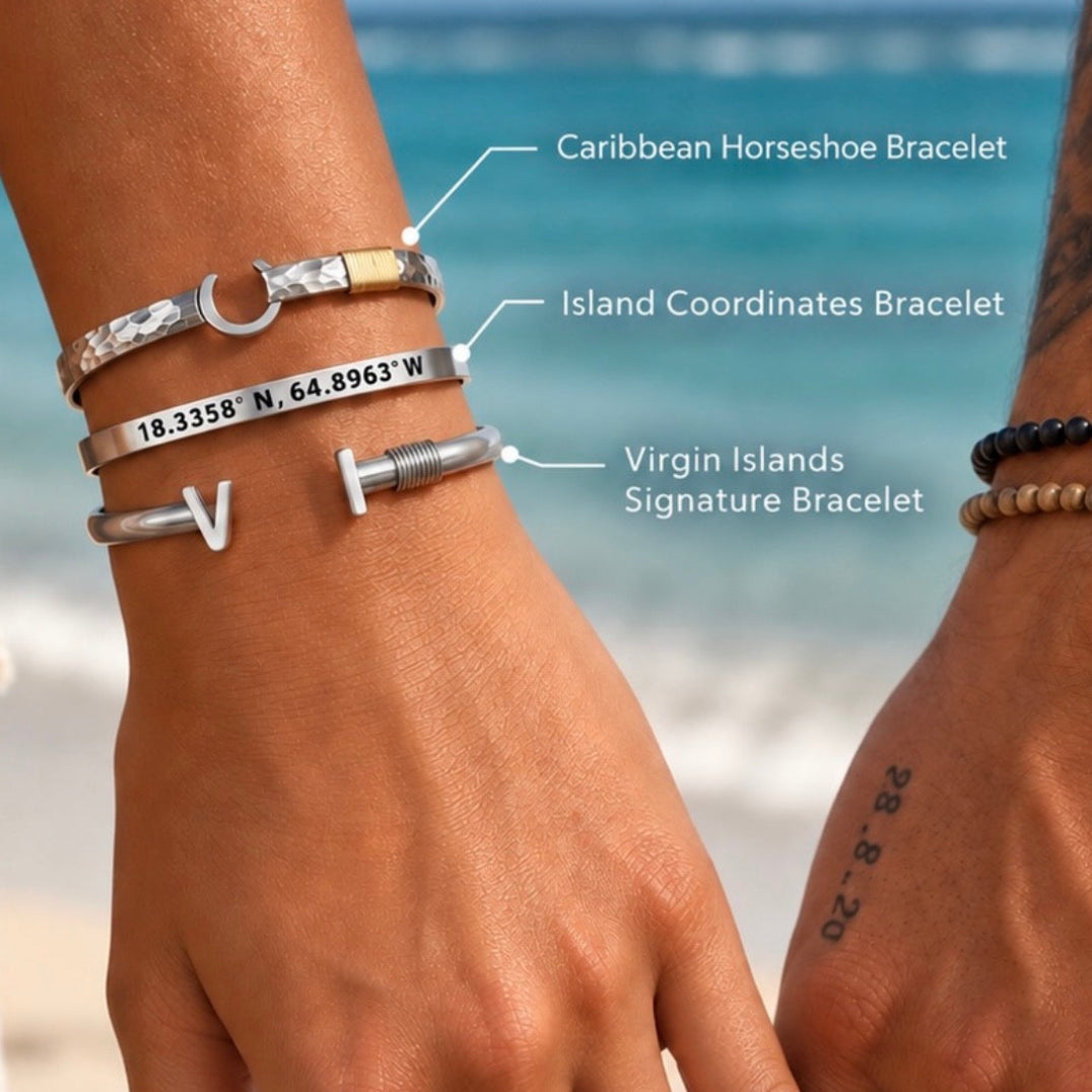 Three silver engraved bracelets worn on a person's wrist, with hand labels pointing to each one: Caribbean Horseshoe Bracelet, Island Coordinates Bracelet, and Virgin Islands Signature Bracelet. The background shows a beach and ocean.
